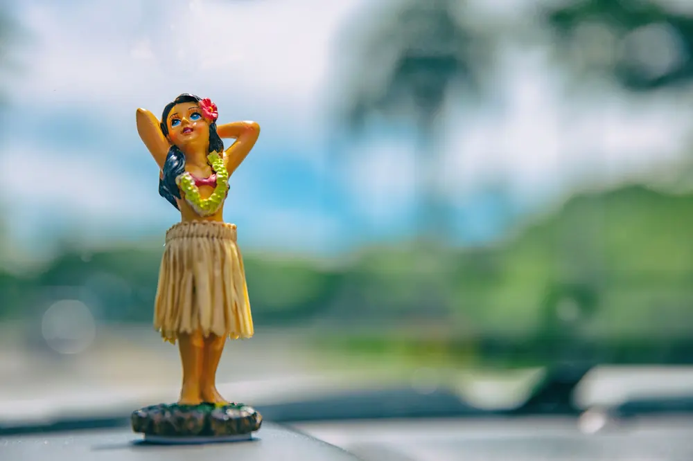 Hula dancer figurine on a rental car dashboard with a blurred Hawaiian landscape behind